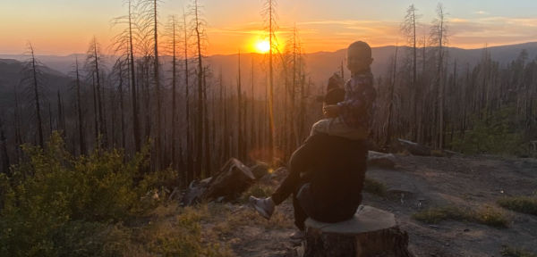 산 중턱에서 아이를 목에 태우고 나무 밑동에 앉아 노을을 바라보고 있는 사진
